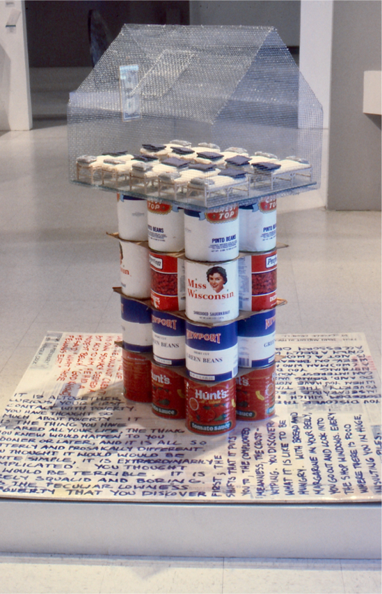 Establishing shot: wire house on top of stacked jumbo cans of food on base with quotes.