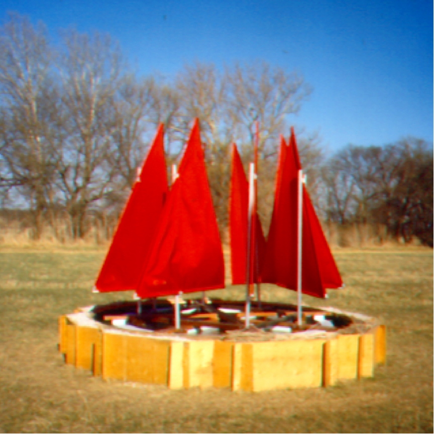 Wheel with 8 red sails floating in an octagonal pool made of plywood set in a large field.
