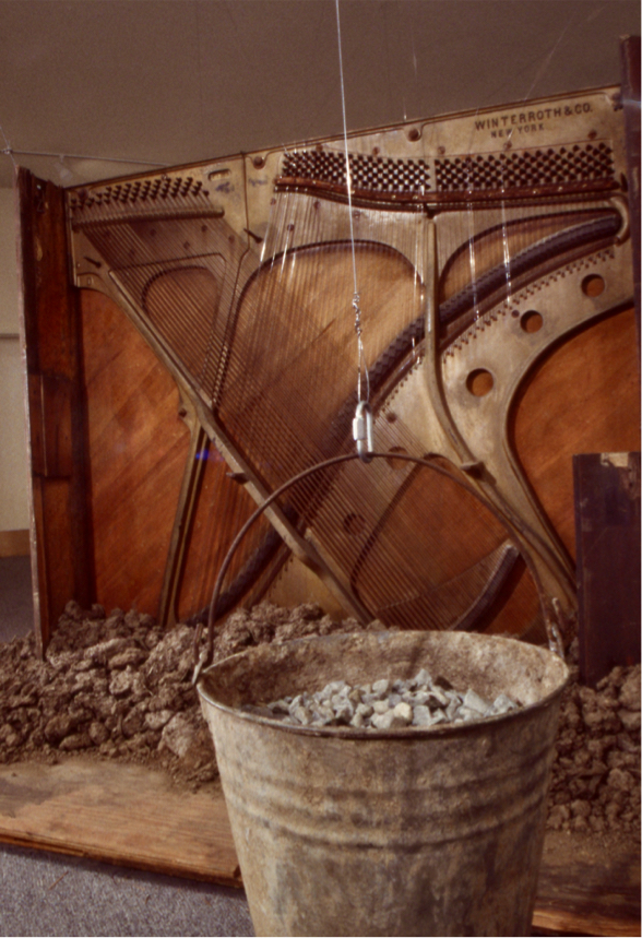 Piano carcass showing exposed harp, suspended metal bucket filled with rocks in foreground.